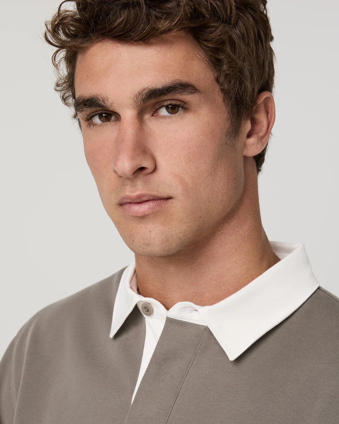 Close-up of a man wearing a taupe knit polo sweater with a contrasting white collar, set against a plain light gray background.