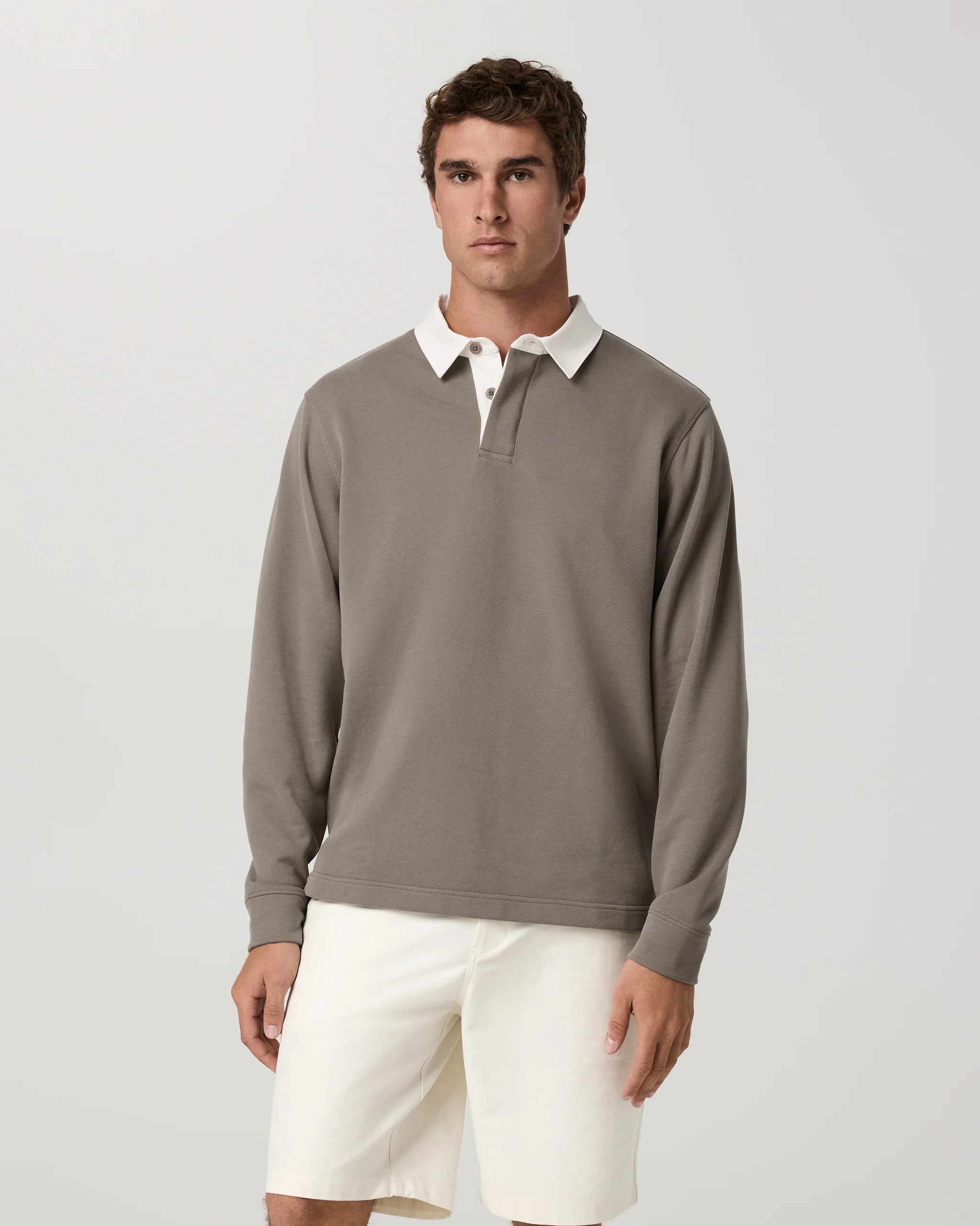 A man wearing a taupe long-sleeve polo shirt with a white collar and white shorts, standing against a light gray studio background.