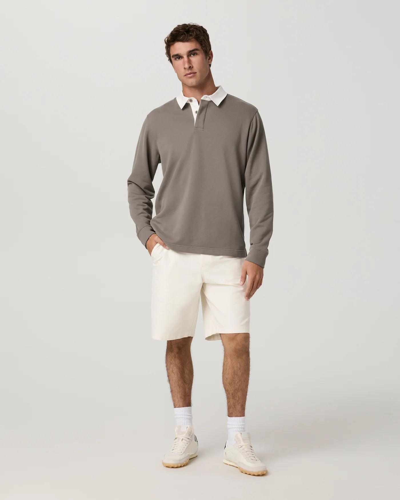 A man modeling a taupe long-sleeve rugby shirt with a white collar, paired with off-white shorts, white socks, and beige sneakers, against a light gray studio background.