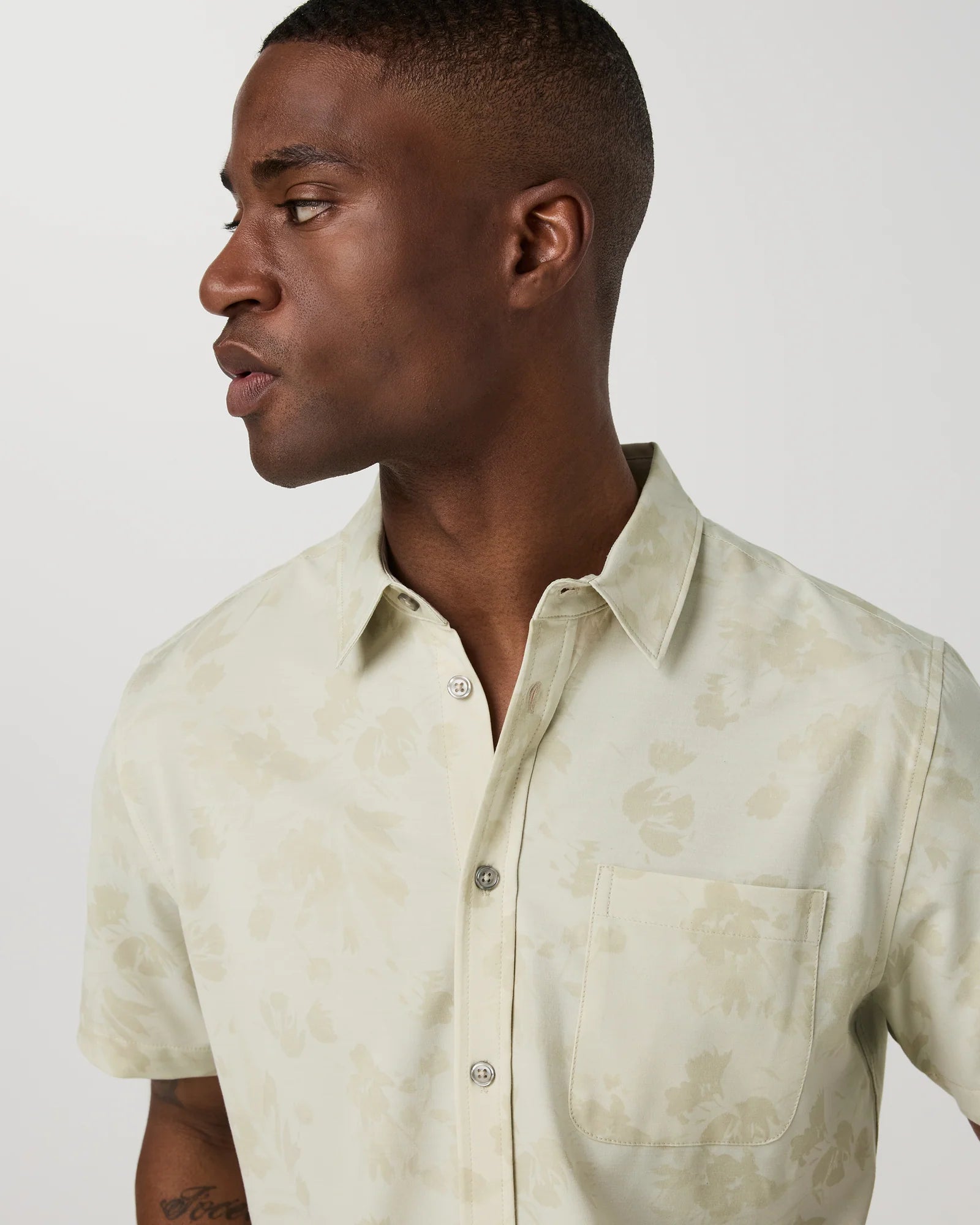 A man in profile wearing a short-sleeve button-up shirt with a light beige and cream floral pattern, photographed against a plain light gray background.
