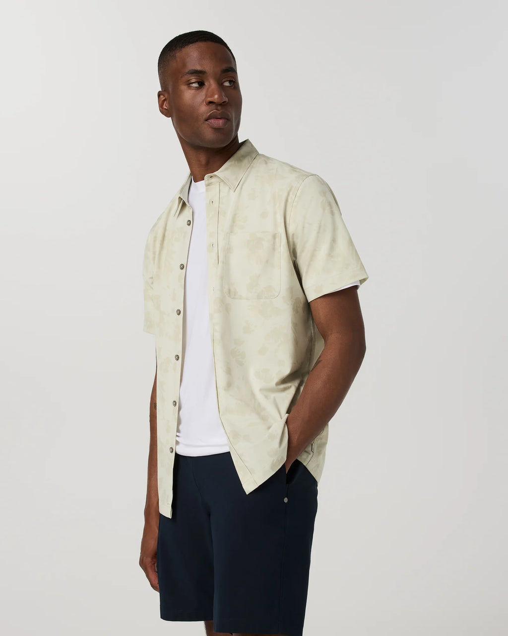 Man wearing a beige short-sleeve shirt over a white t-shirt with navy shorts on a light gray background