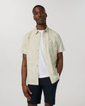 A man wearing a short-sleeve button-up shirt in a light cream color with a subtle tonal pattern, layered over a white t-shirt and dark navy shorts, against a plain light gray background.