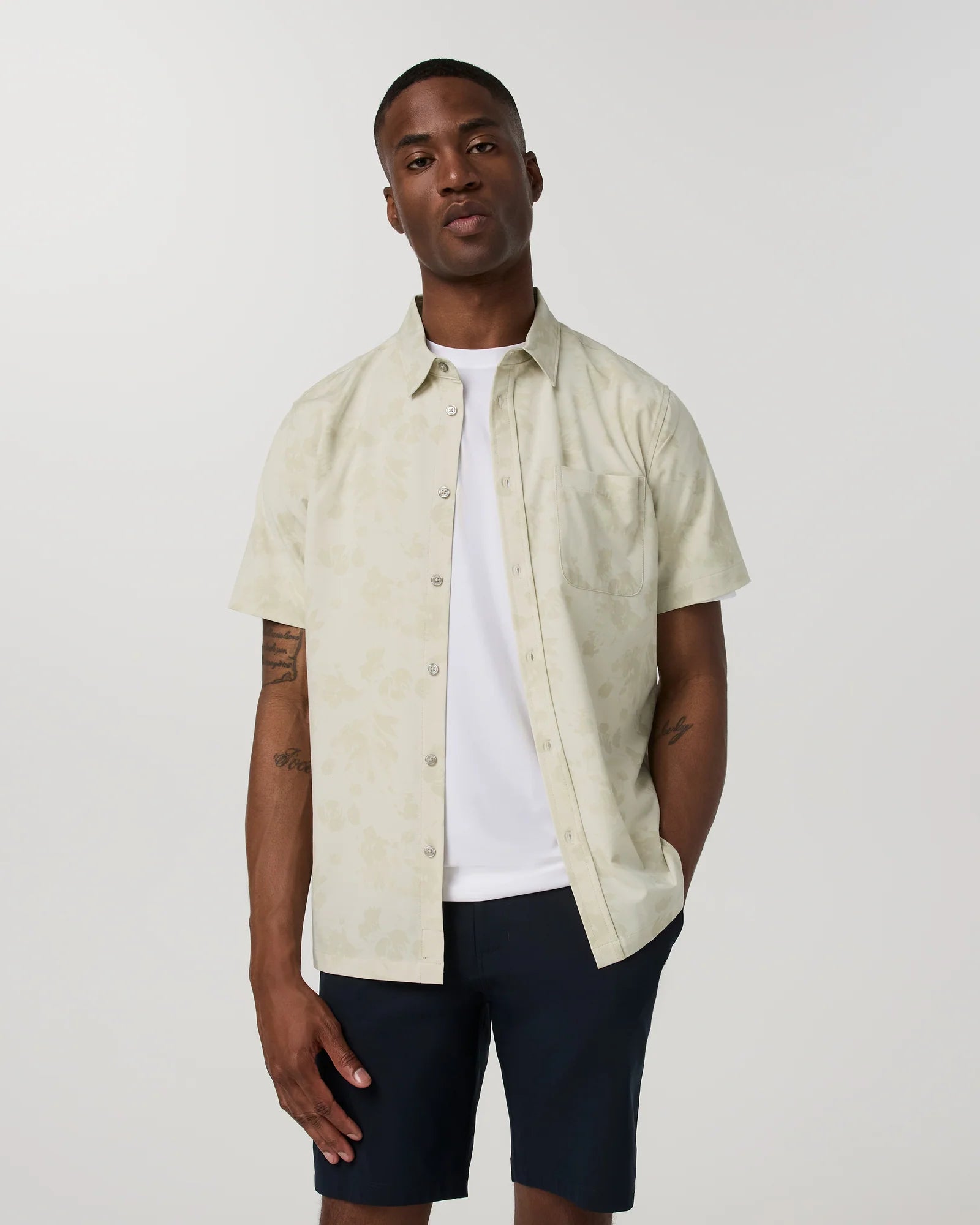 A man wearing a short-sleeve button-up shirt in a light cream color with a subtle tonal pattern, layered over a white t-shirt and dark navy shorts, against a plain light gray background.