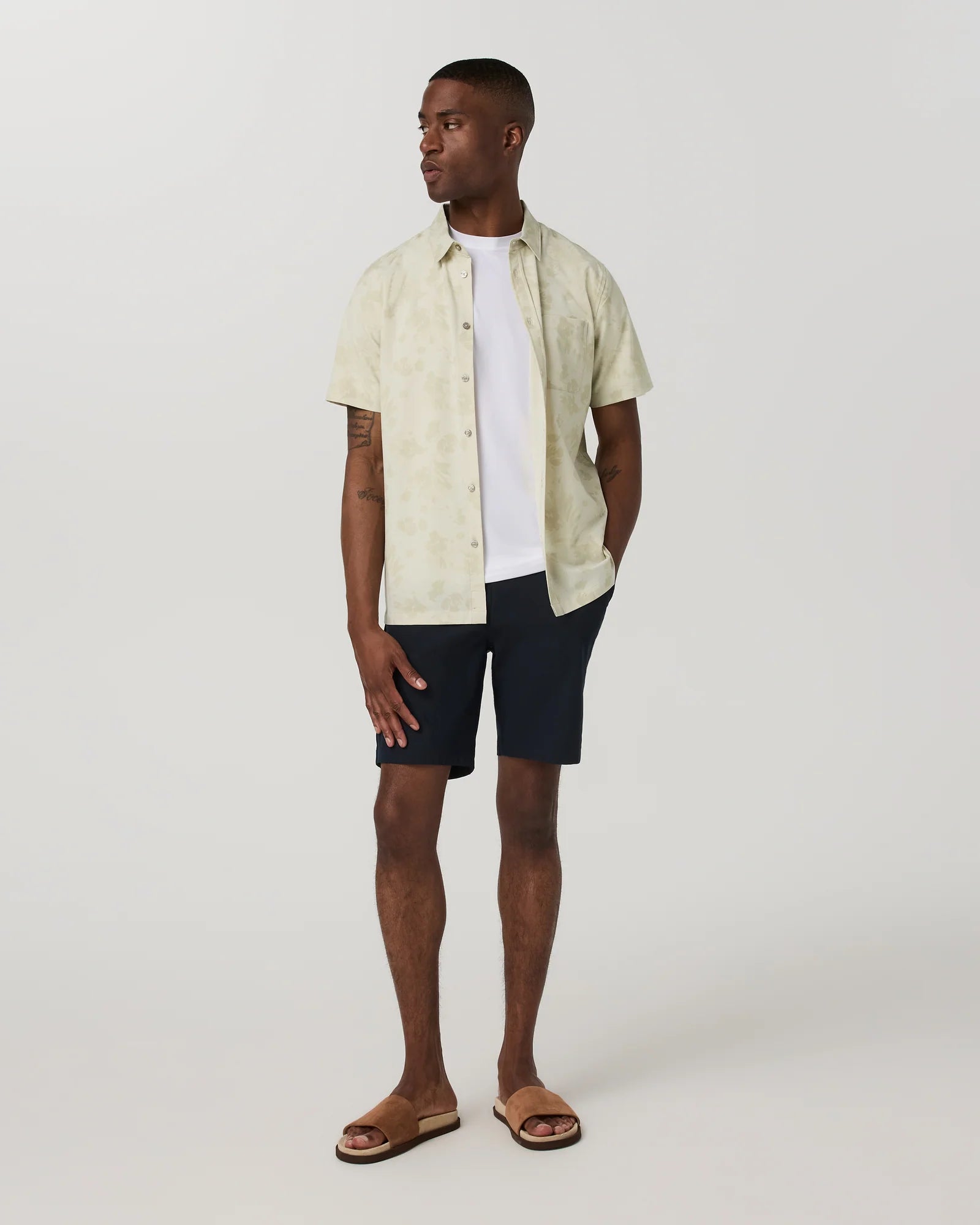 Man wearing a light-colored shirt over a white t-shirt and dark shorts on a plain background