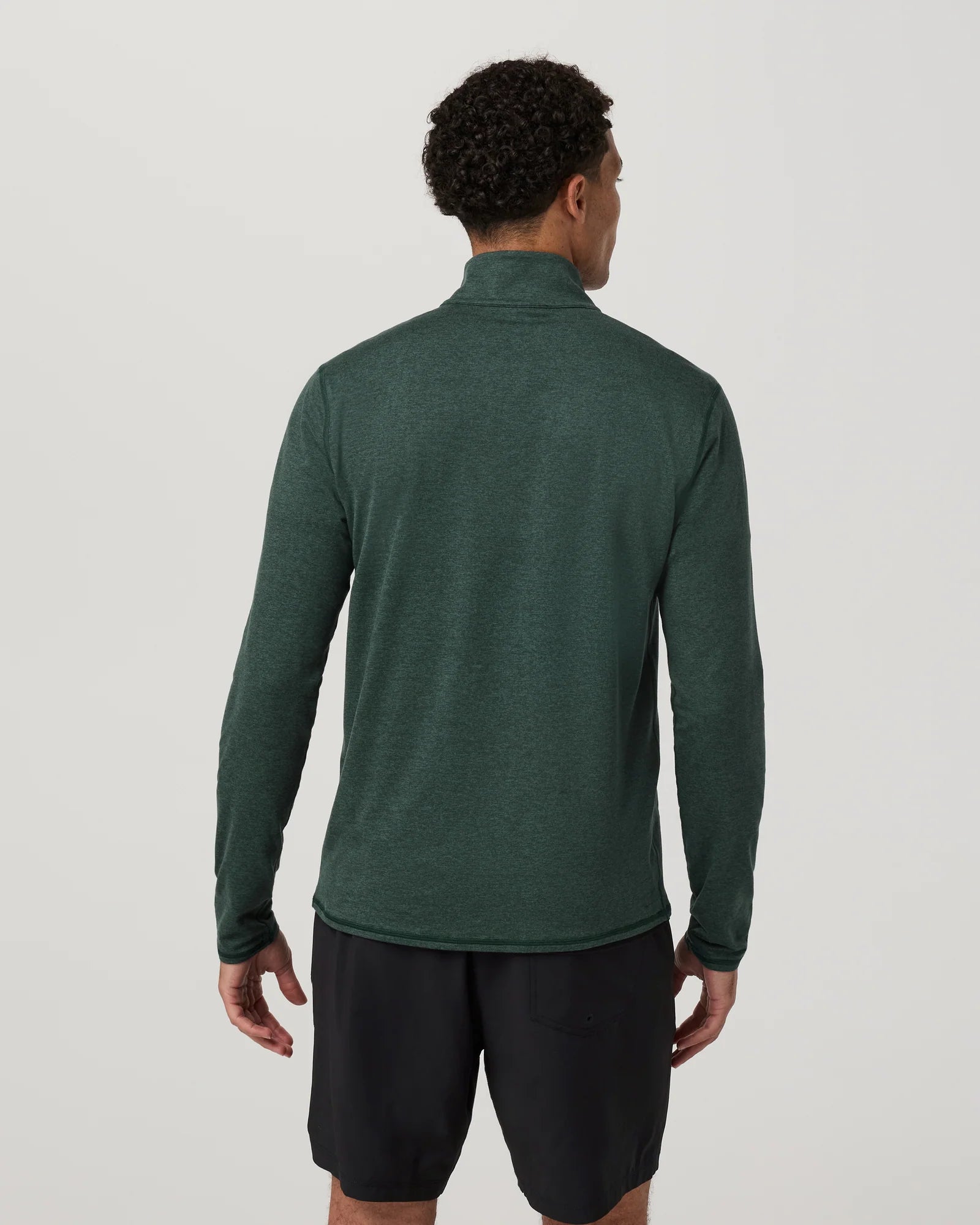 A man viewed from the back wearing a dark green long-sleeve quarter-zip athletic top and black shorts against a light gray studio background.