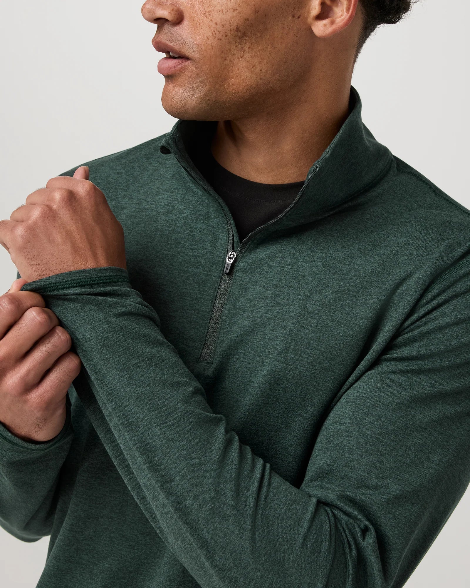 A close-up of a man wearing a dark green quarter-zip long-sleeve shirt over a black undershirt, adjusting the sleeve cuff against a light gray background.
