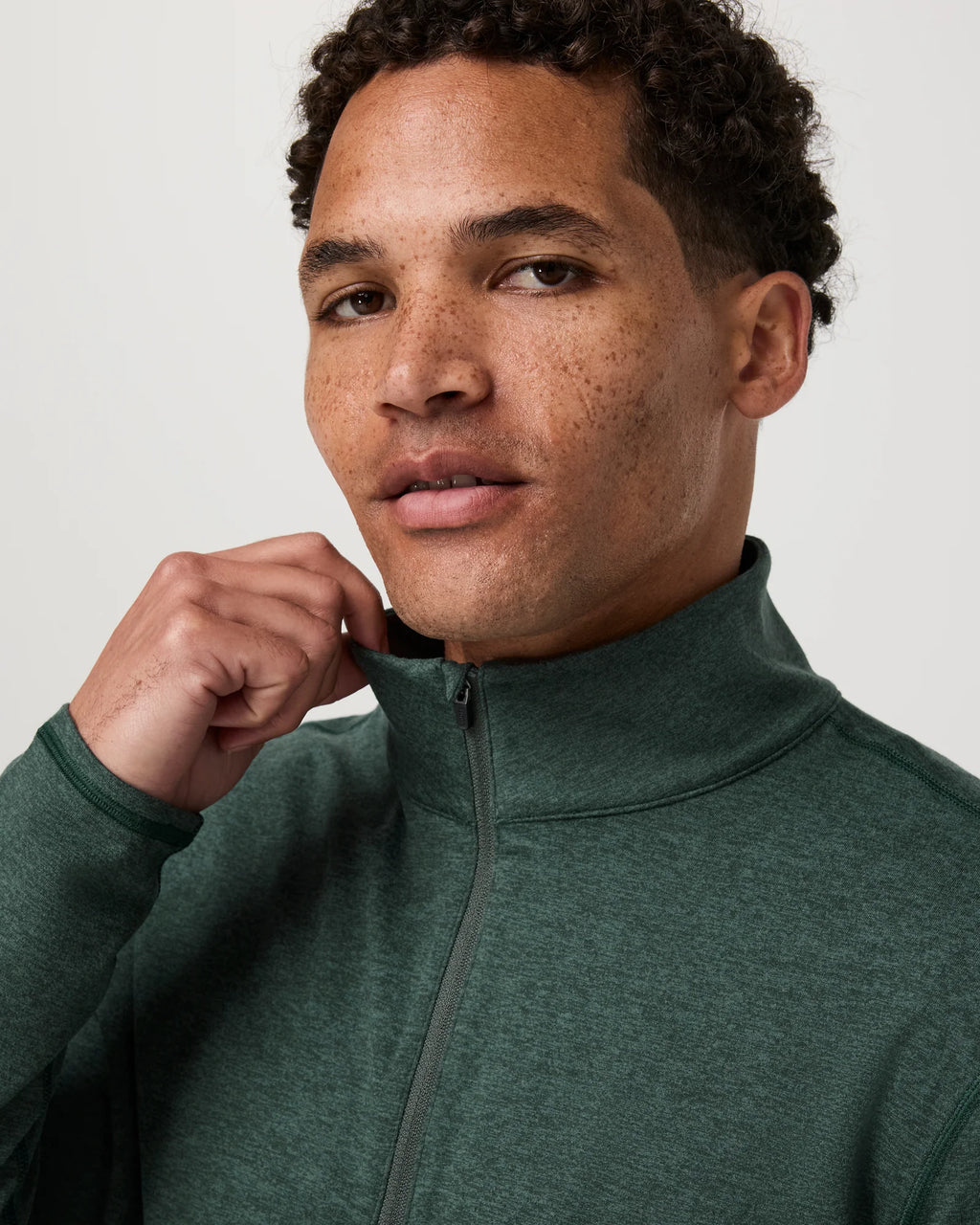 A close-up of a man wearing a dark green heathered quarter-zip pullover jacket, adjusting the collar with one hand against a plain light gray background.