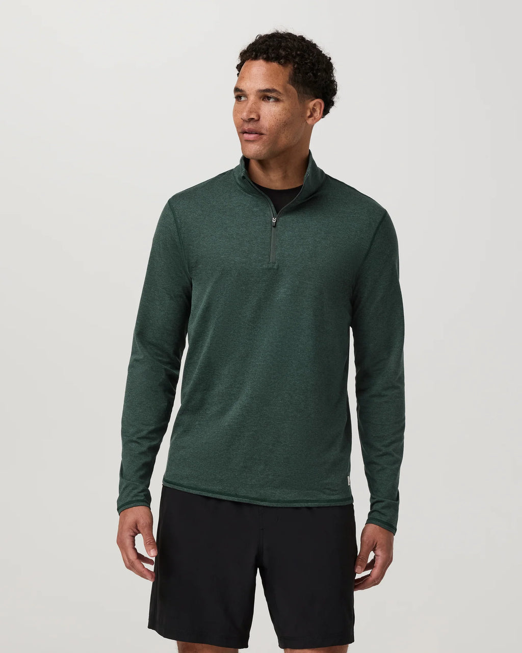 A man wearing a dark green long-sleeve quarter-zip athletic pullover and black athletic shorts, standing against a light gray studio background.
