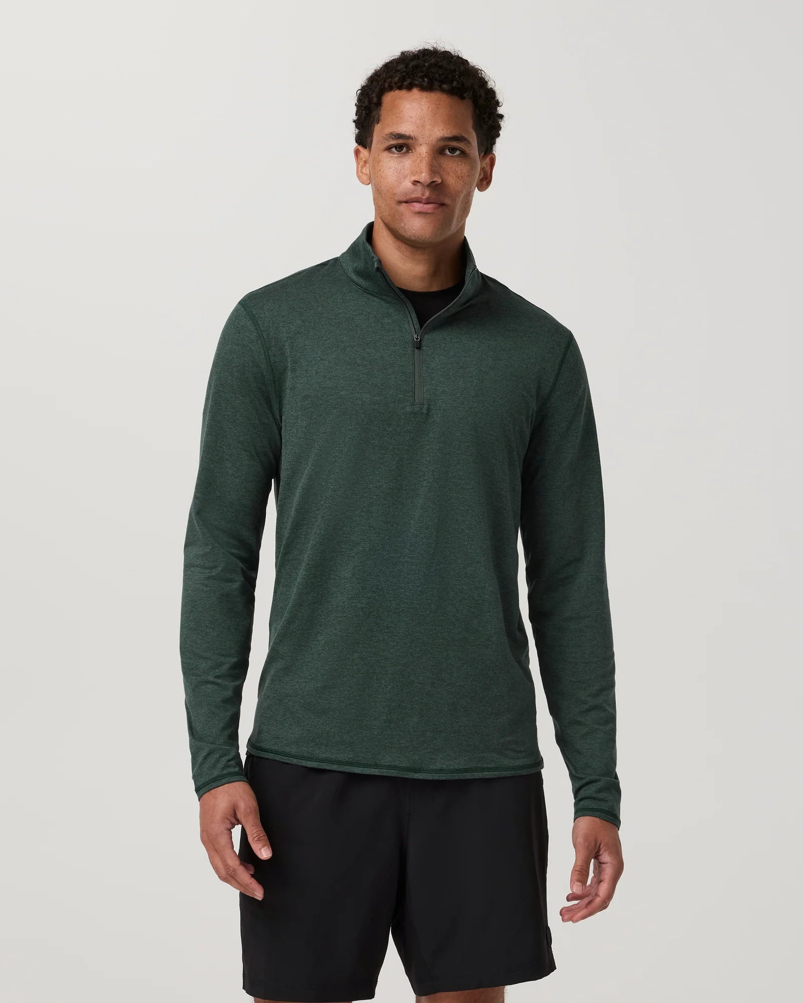 A man wearing a dark green long-sleeve quarter-zip athletic pullover and black shorts, standing against a light gray studio background.