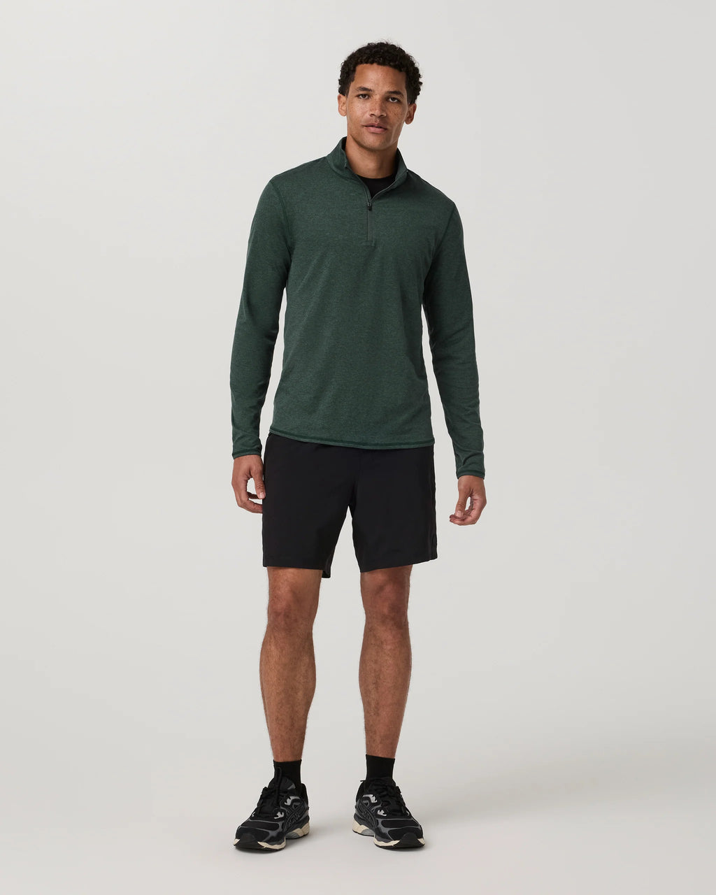 A man modeling a dark green long-sleeve quarter-zip athletic pullover, paired with black athletic shorts, black socks, and black running shoes, against a light gray studio background.