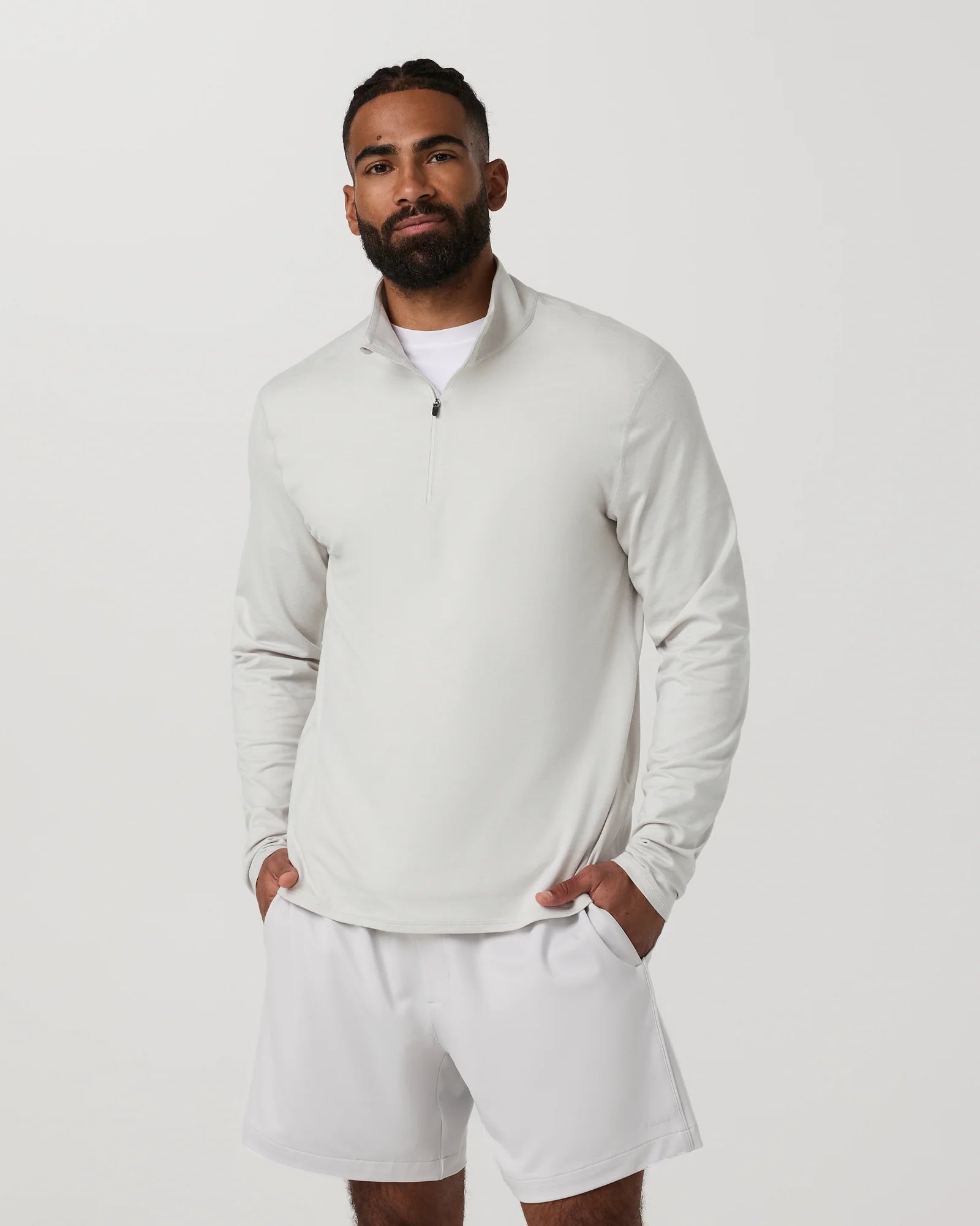 A man wearing a light gray quarter-zip long-sleeve shirt and matching light gray shorts, standing against a plain light gray studio background.