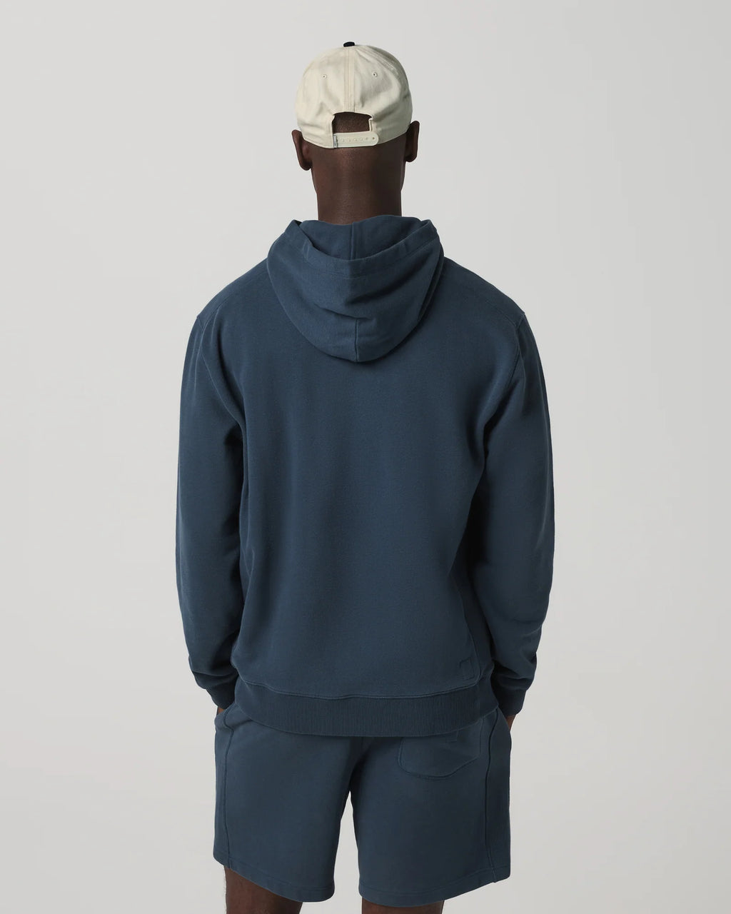 Person wearing a navy blue hoodie and shorts set with a beige cap on a light gray background