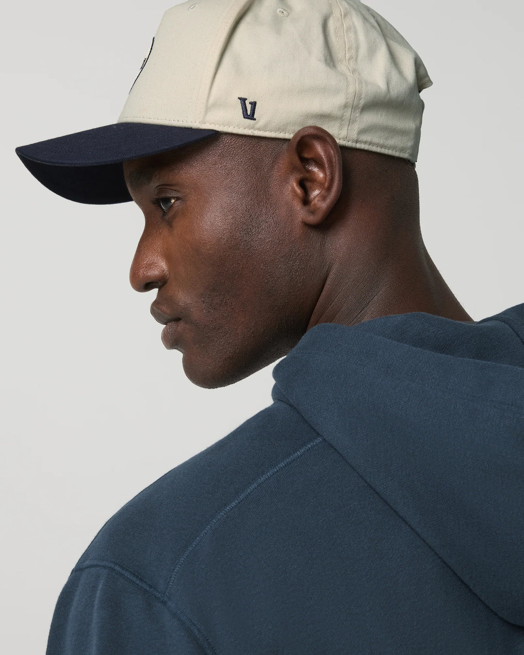 Man wearing a beige cap and navy hoodie on a plain background