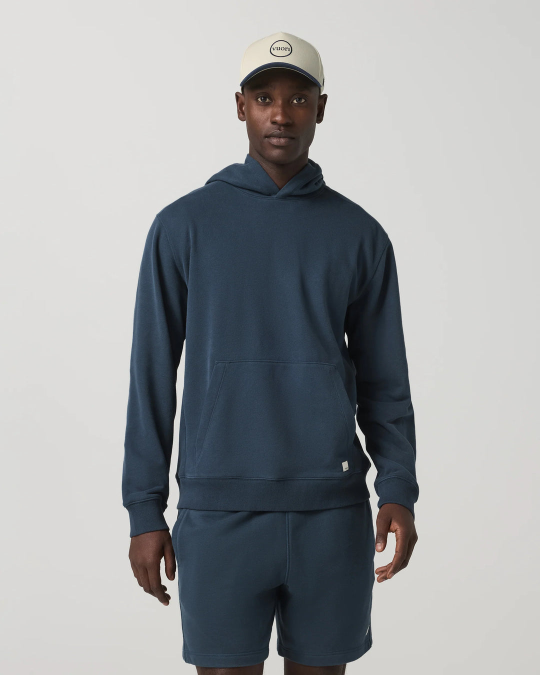 Man wearing a navy hoodie and shorts with a beige cap on a light gray background