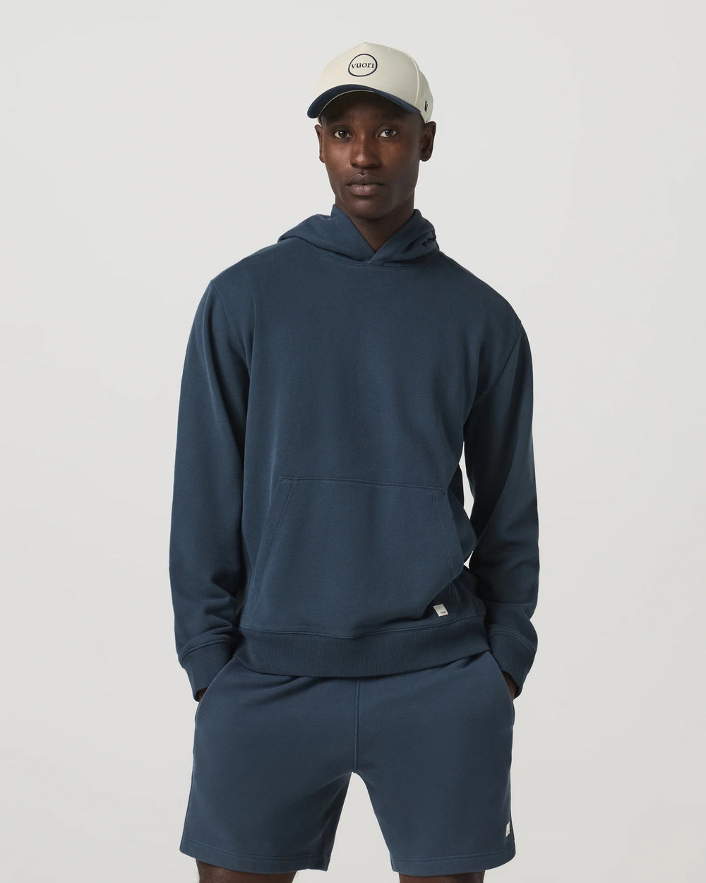 Man wearing a navy hoodie and shorts set with a cap on a light gray background