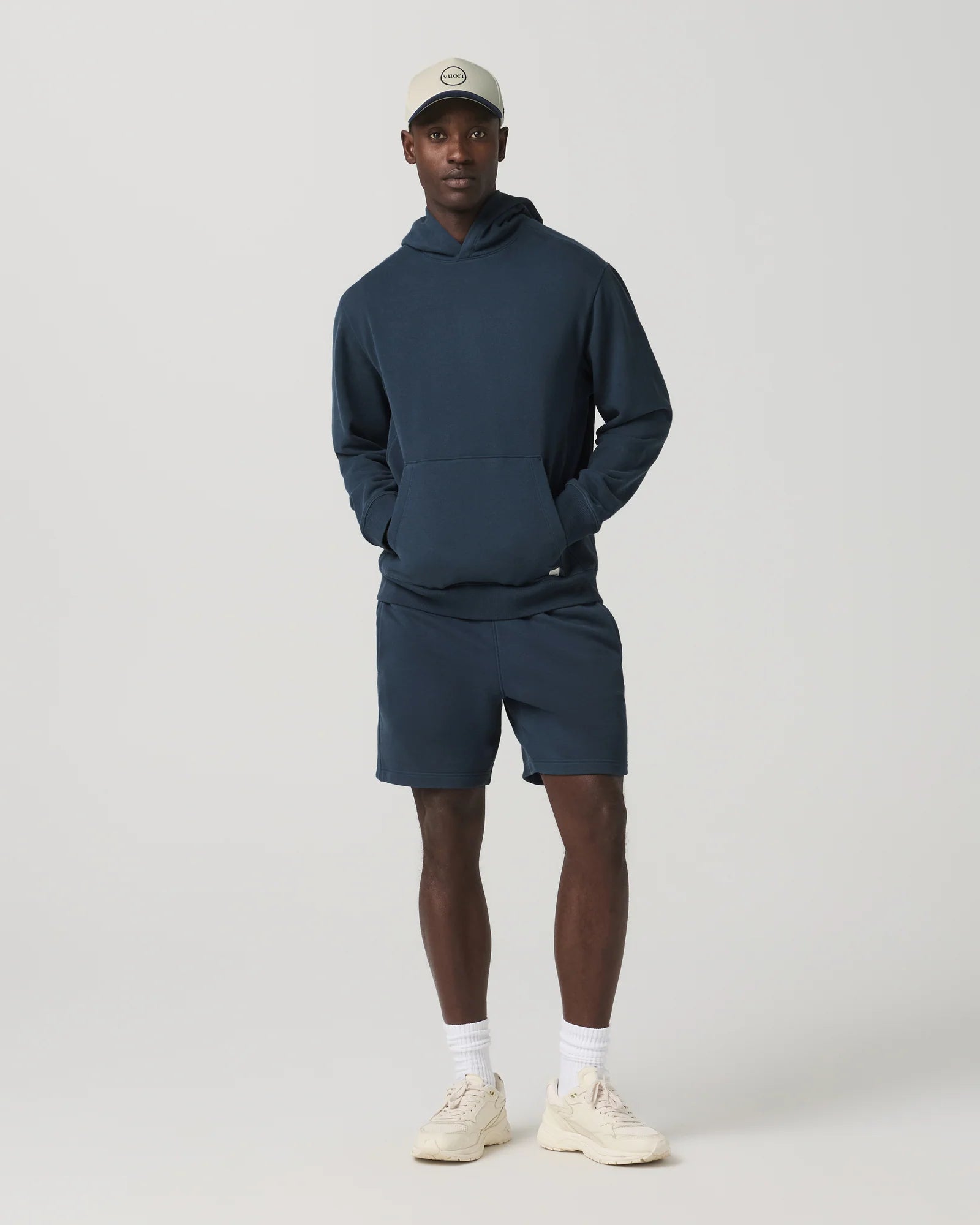 Man wearing a navy hoodie and shorts with a white cap on a light gray background