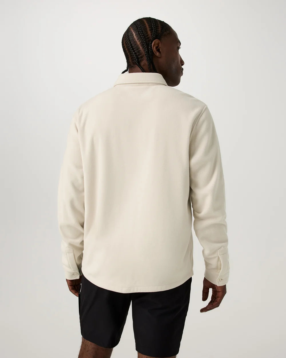 A man viewed from the back wearing a cream-colored long-sleeve button-down shirt and black shorts, standing against a light gray studio background.