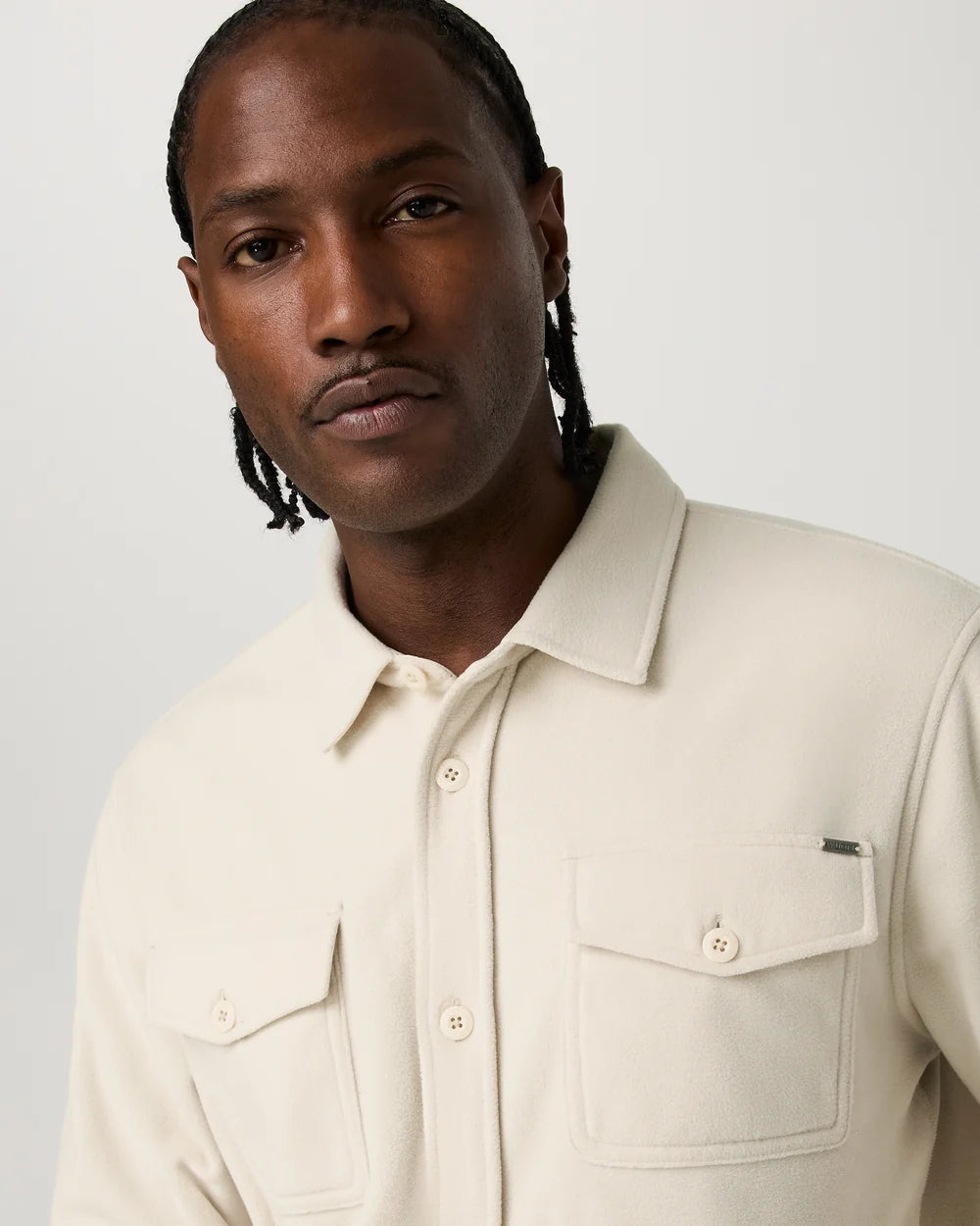 A man wearing a light cream-colored button-up overshirt over a white t-shirt and black shorts, standing against a solid light gray background.