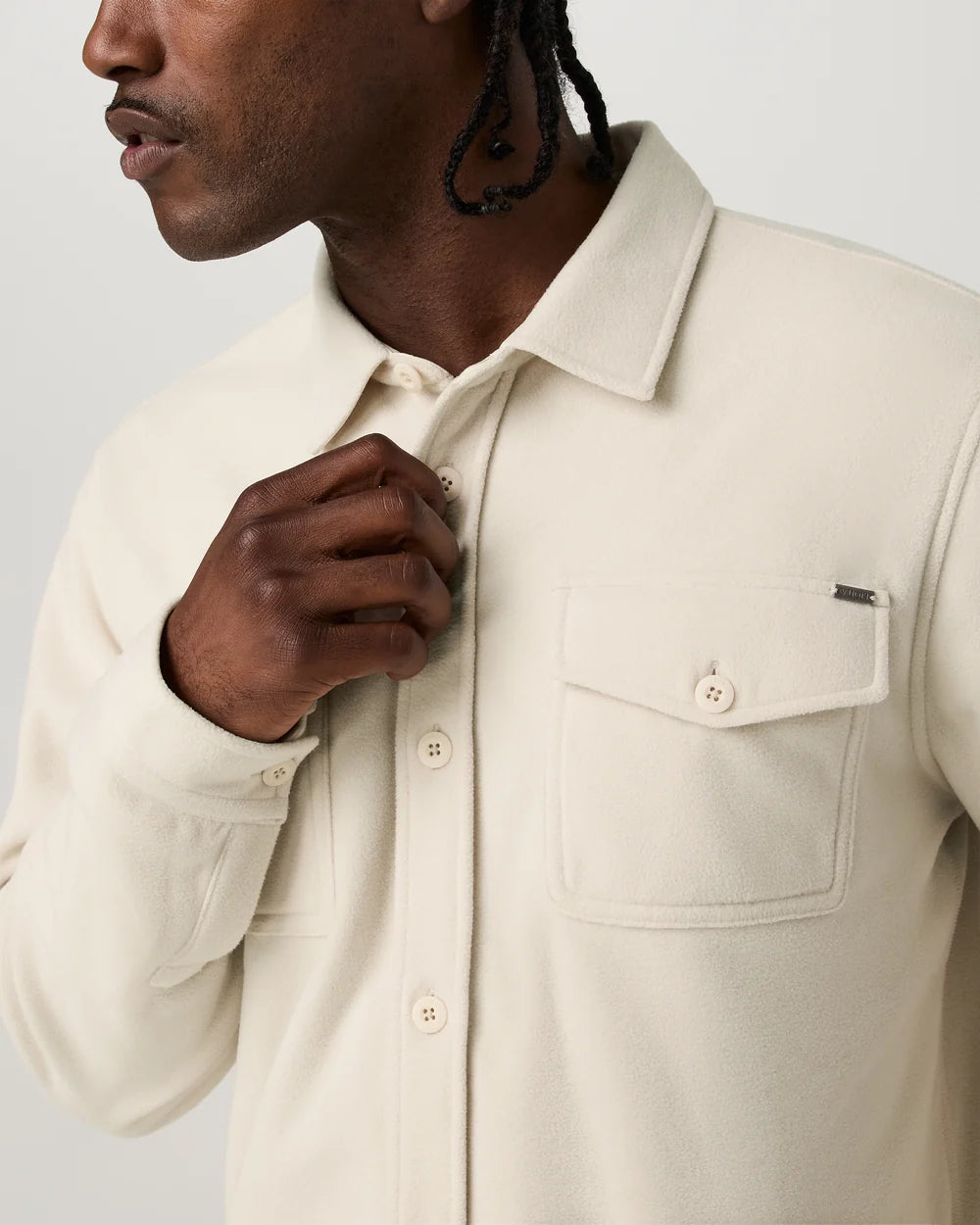 A close-up of a man wearing a cream-colored long-sleeve button-up shirt with a chest pocket and button details, adjusting the collar and placket.