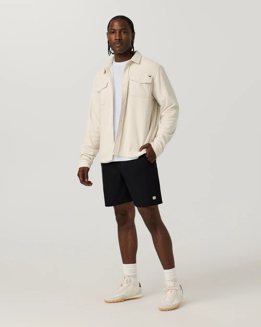 A male model stands against a light gray studio background wearing a cream-colored overshirt, black athletic shorts, a white t-shirt, white socks, and off-white sneakers.