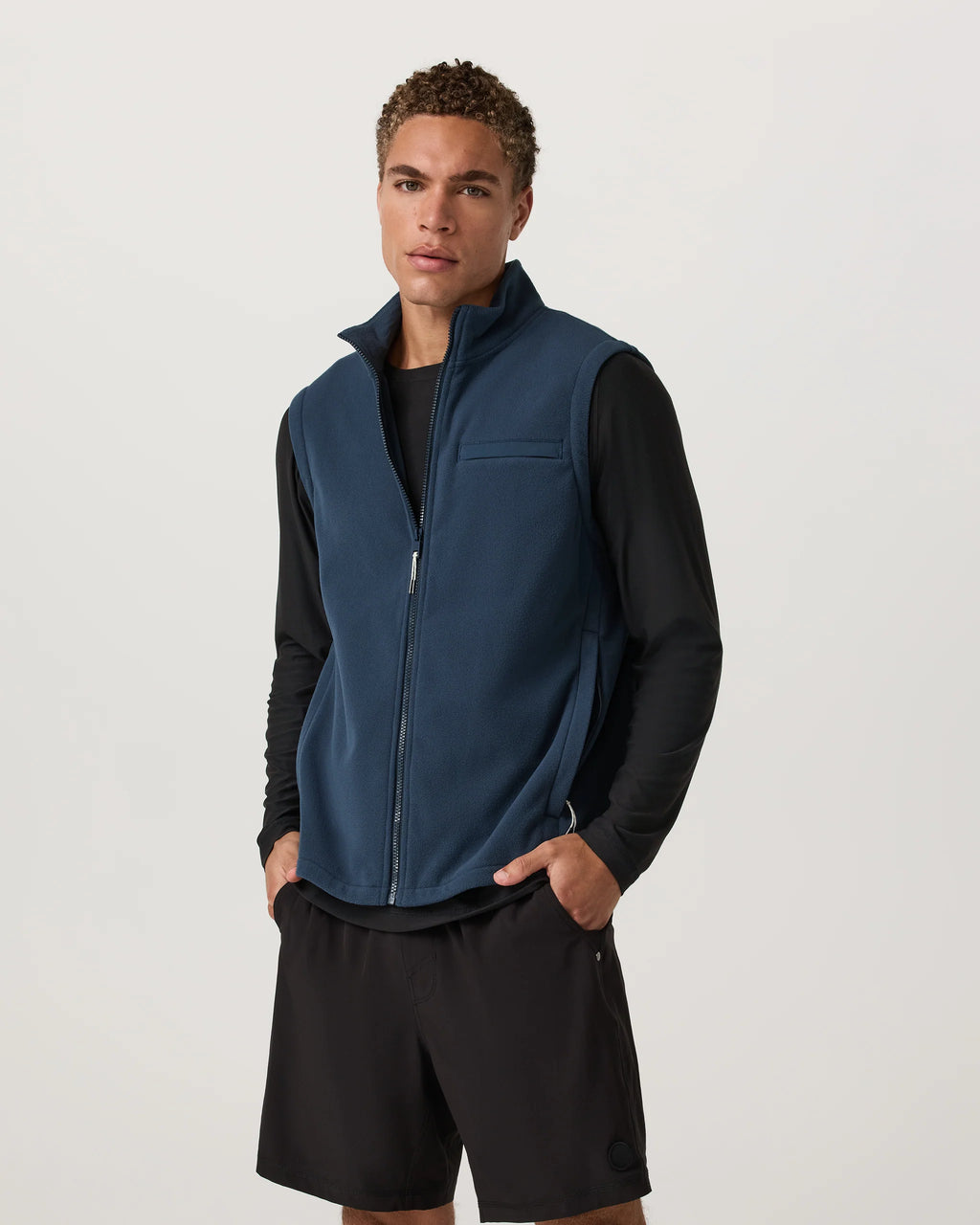 Man wearing a blue vest over a black shirt and shorts on a white background