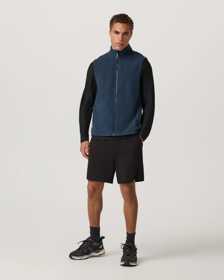 Man wearing a navy vest over a black long-sleeve shirt and black shorts on a white background