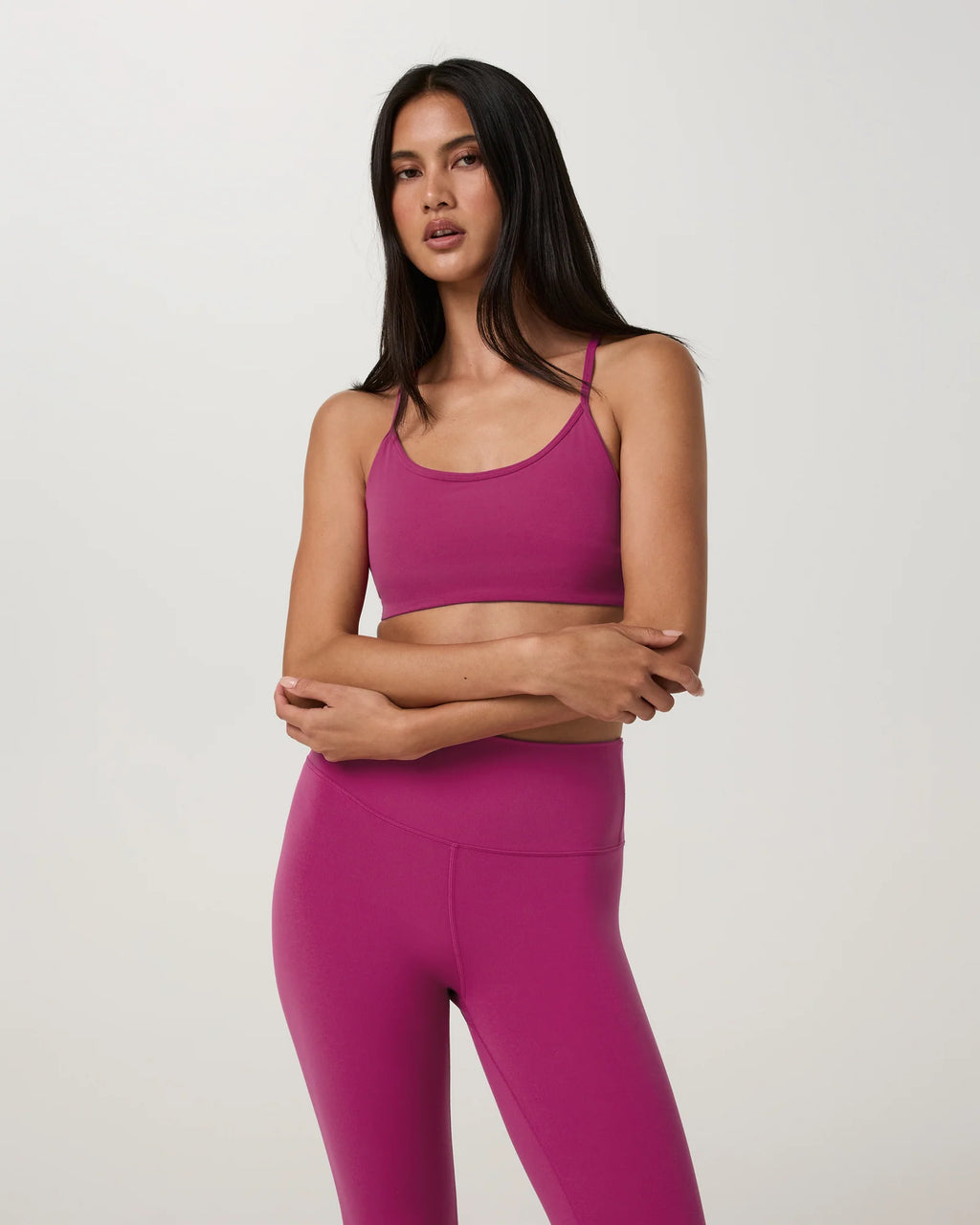 A woman modeling a matching magenta activewear set consisting of a sports bra and high-waisted leggings, photographed against a neutral light gray studio background.