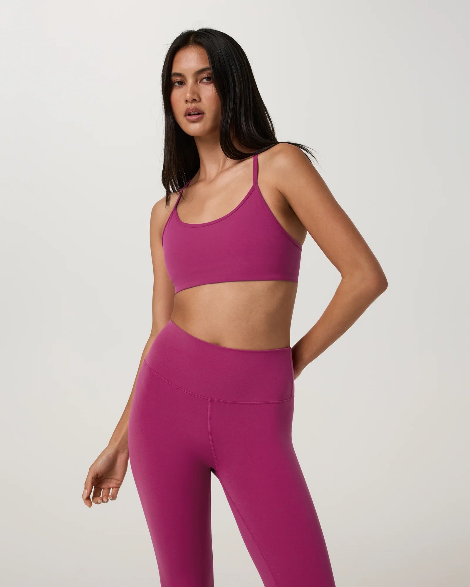 A woman modeling a matching magenta activewear set consisting of a sports bra and high-waisted leggings, photographed against a neutral light gray studio background.
