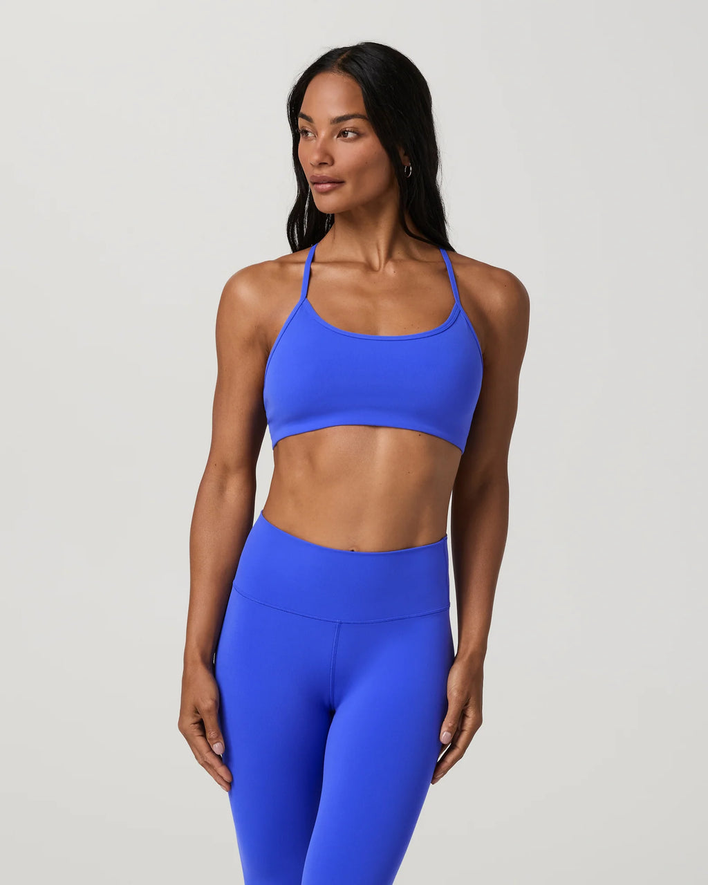 A woman modeling a bright royal blue activewear set consisting of a sports bra and high-waisted leggings, photographed against a solid light gray studio background.