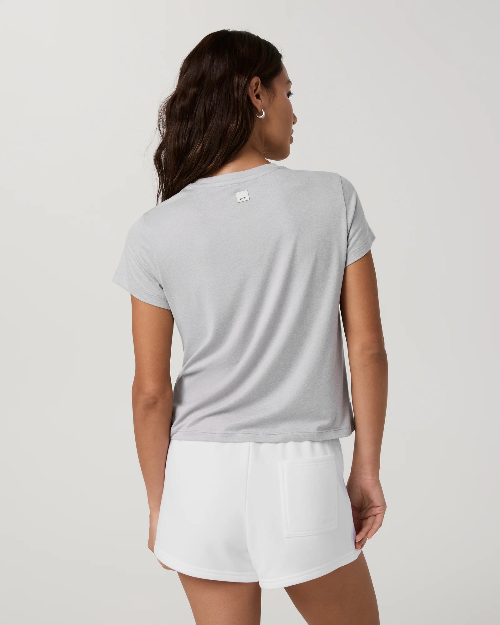 A woman shown from the back wearing a light gray short-sleeve t-shirt and white shorts against a plain light gray studio background.