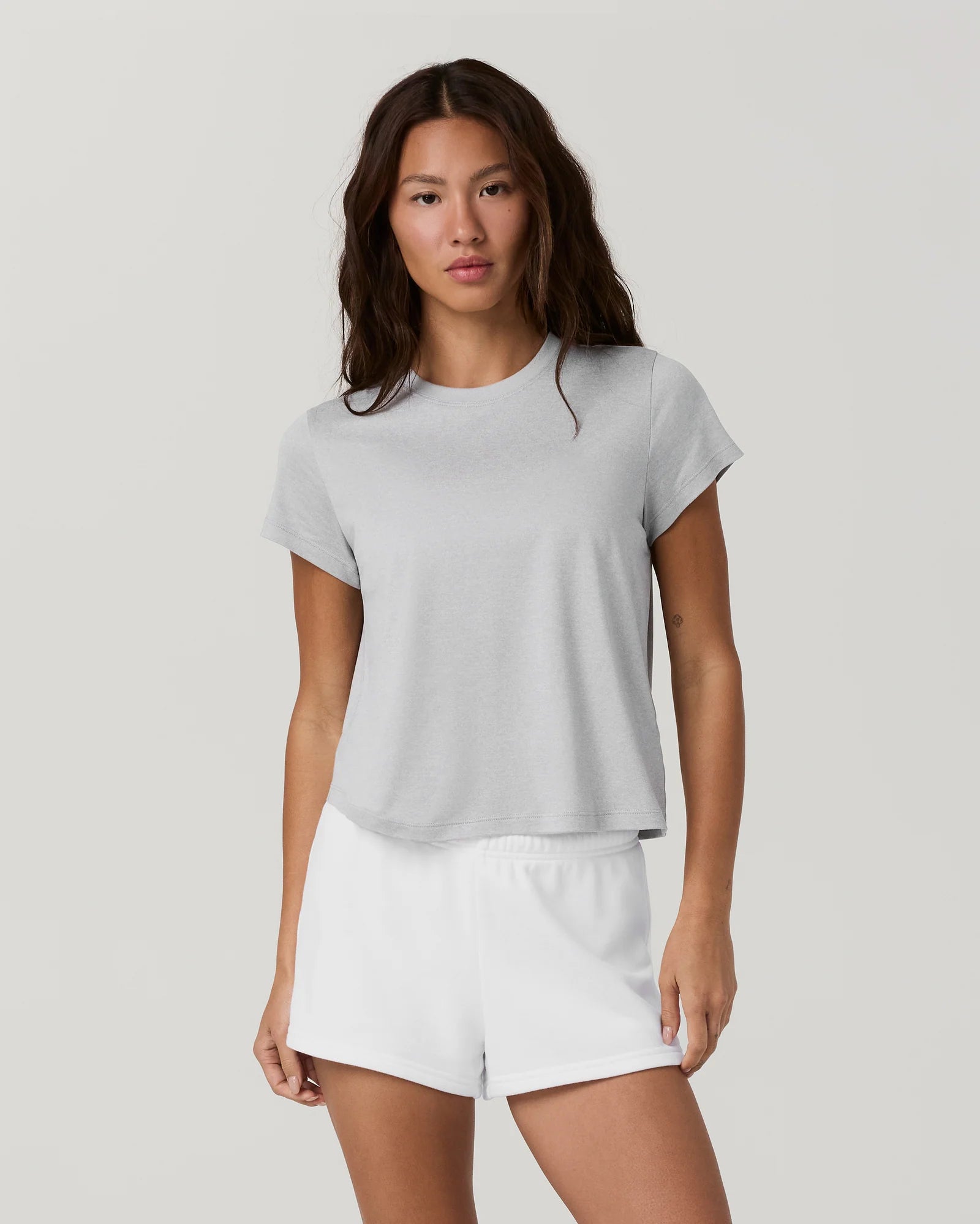 A woman modeling a light gray short-sleeve cropped t-shirt and white athletic shorts against a solid light gray studio background.