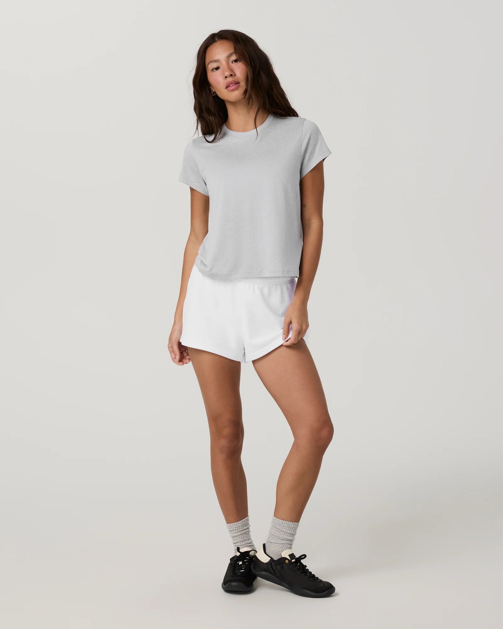 A woman modeling a light gray short-sleeve t-shirt and white athletic shorts against a plain light gray studio background.