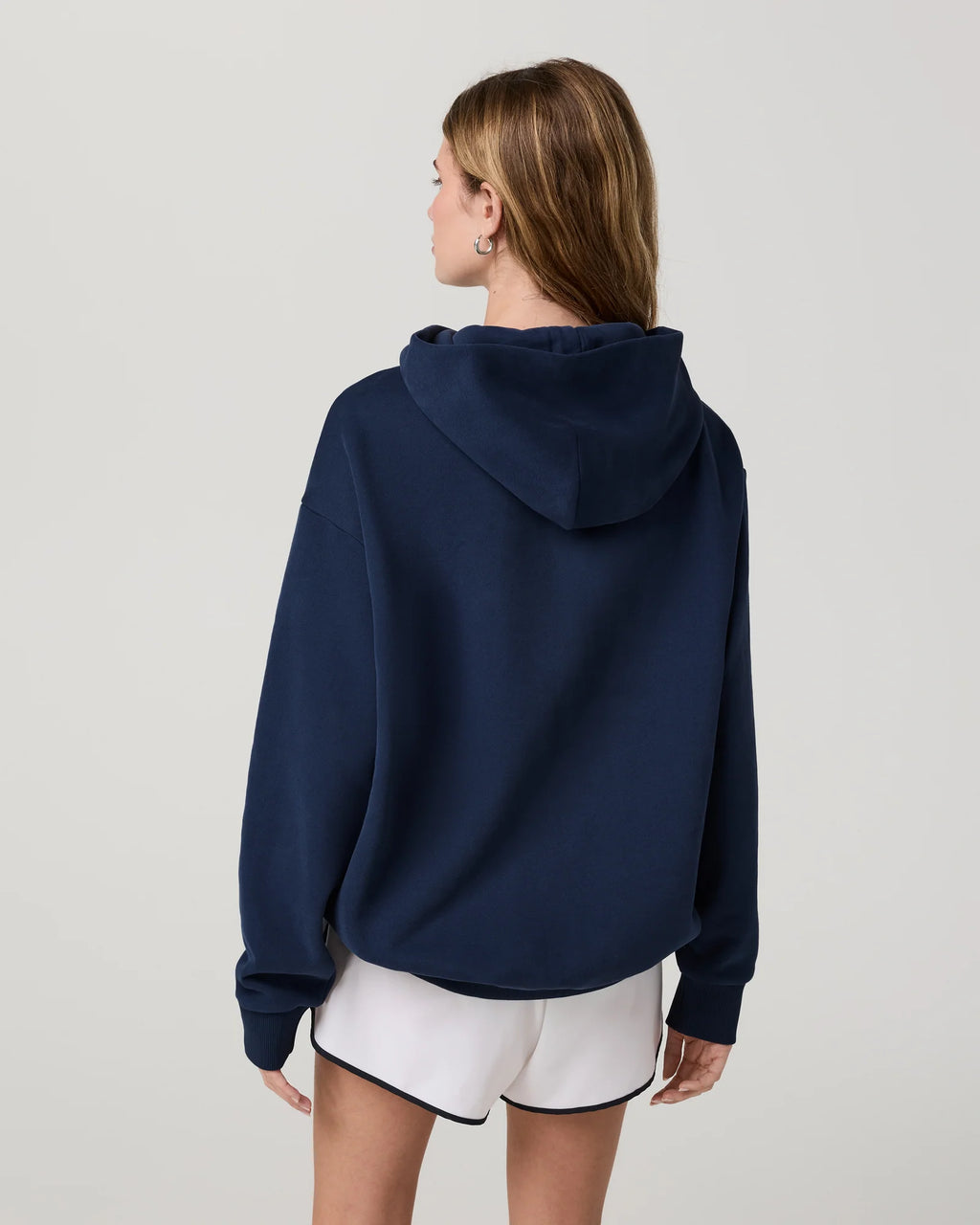 A woman shown from the back wearing a navy blue cropped hoodie and white shorts with black trim, photographed against a plain light gray studio background.