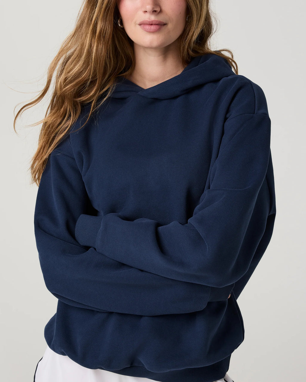A woman wearing an oversized navy blue hoodie with her arms crossed, shown from the chest up against a light gray studio background.