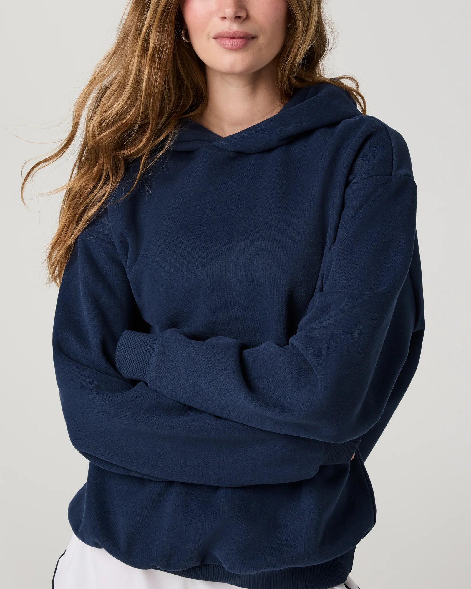 A woman wearing an oversized navy blue hoodie with her arms crossed, shown from the chest up against a light gray studio background.
