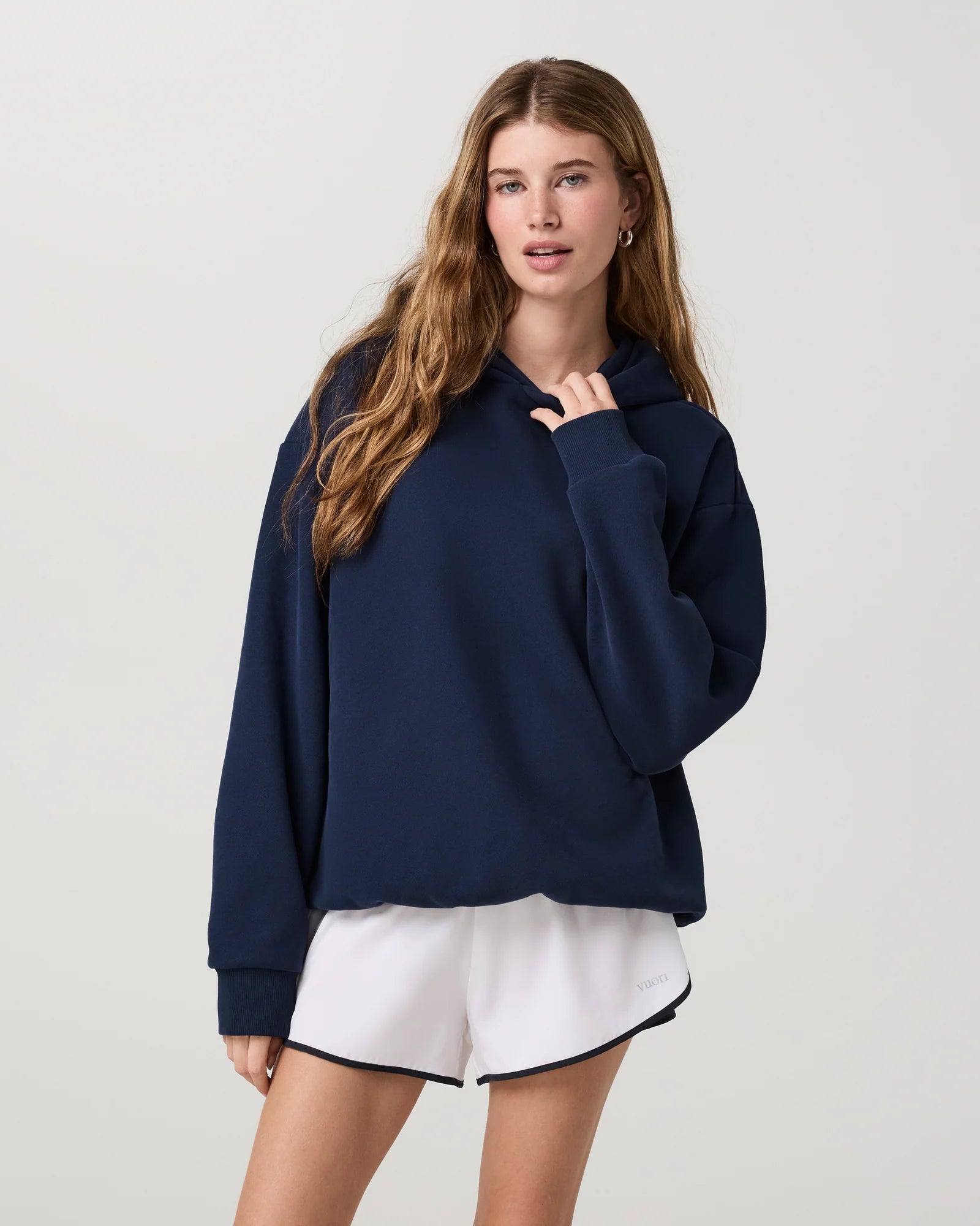 A woman modeling an oversized navy blue hoodie with white athletic shorts against a light gray studio background.