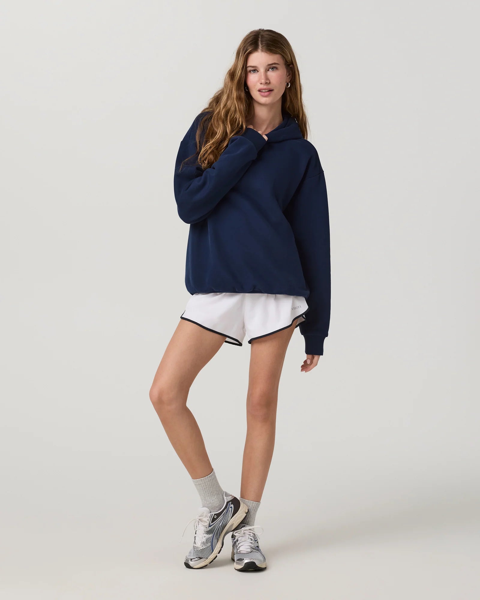 A woman modeling a navy blue oversized hoodie, white athletic shorts with black trim, white socks, and gray running shoes against a light gray studio background.
