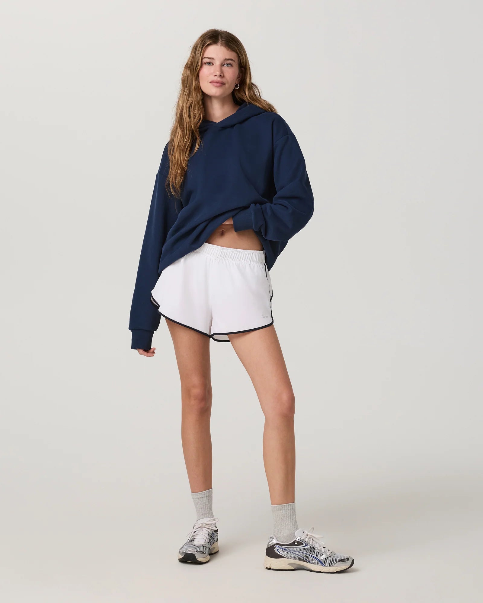 A woman modeling a navy blue oversized hoodie, white athletic shorts with black trim, gray socks, and gray running shoes against a light gray studio background.