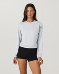 A woman modeling a light gray heather crewneck sweatshirt paired with black athletic shorts, photographed against a solid light gray studio background.