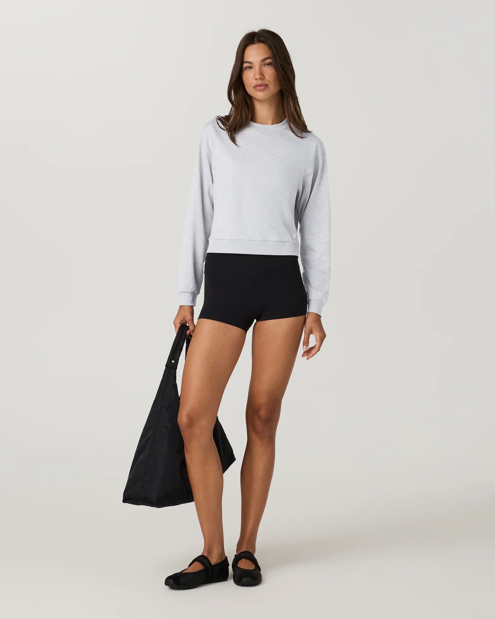 A model stands against a light gray studio background wearing a light gray long-sleeve crewneck sweatshirt, black fitted shorts, black flats, and holding a black tote bag.