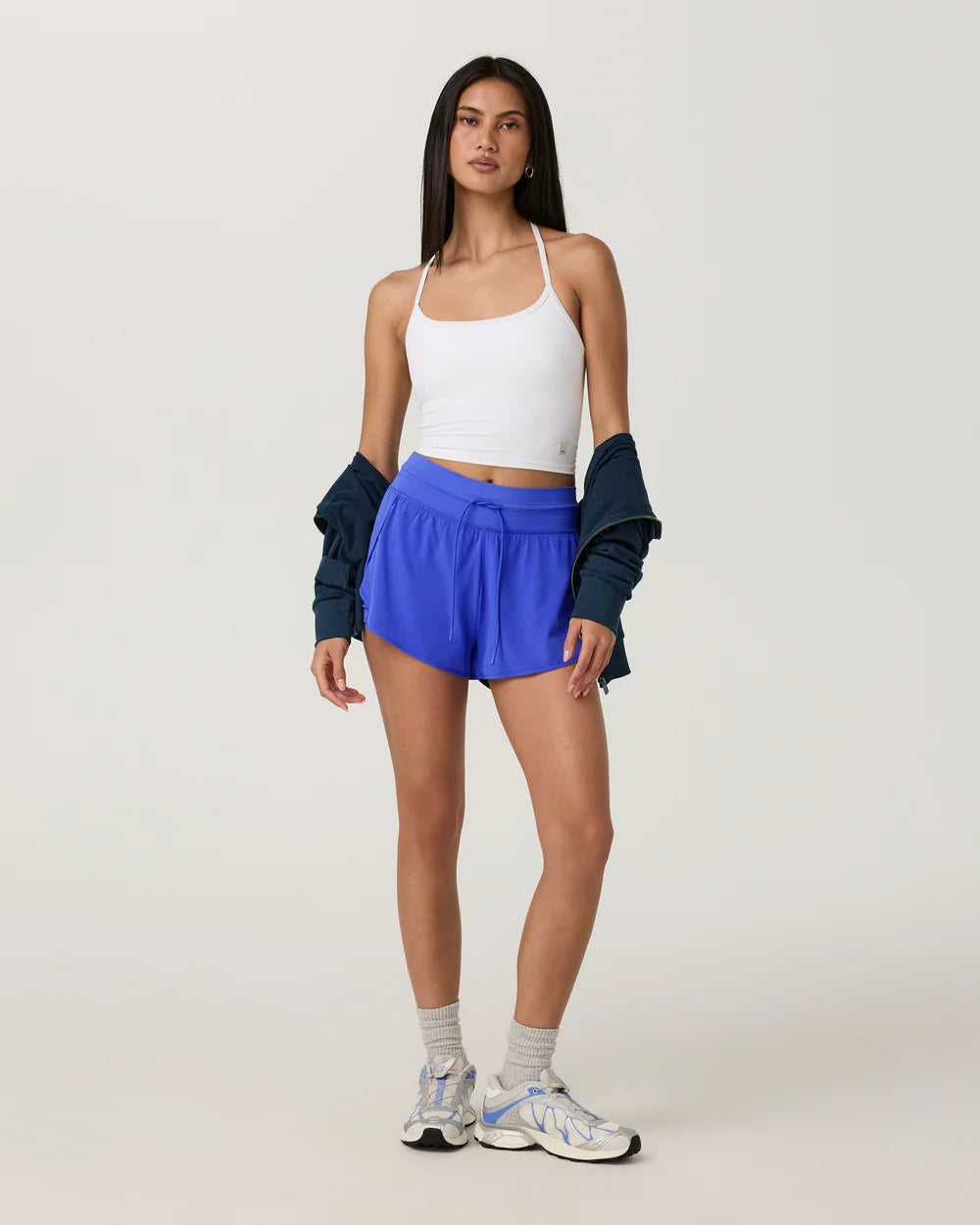 A woman modeling a white ribbed tank top, bright blue athletic shorts, a dark navy cardigan draped off her shoulders, white socks, and white and blue sneakers, set against a plain light gray studio background.