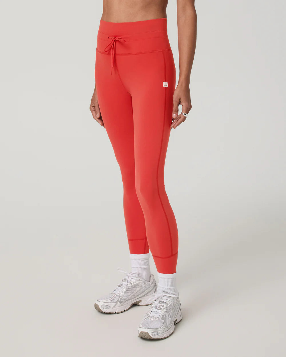 A model wearing bright coral-red high-waisted athletic leggings with a drawstring waist, paired with white socks and white sneakers, shown from the waist down against a light gray studio background.