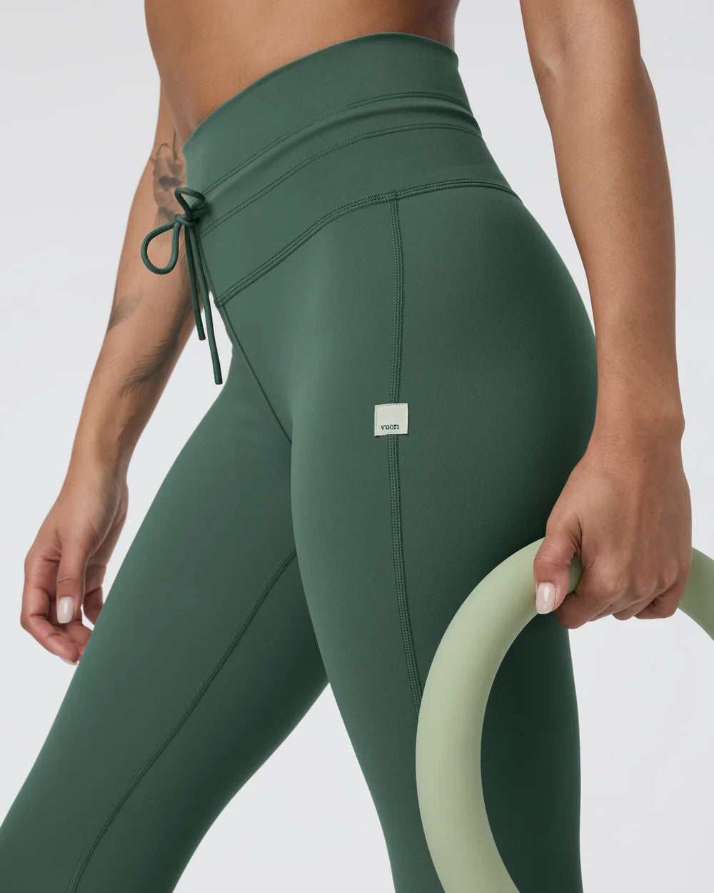 Person wearing green leggings with a small logo, holding a green exercise ring on a light gray background
