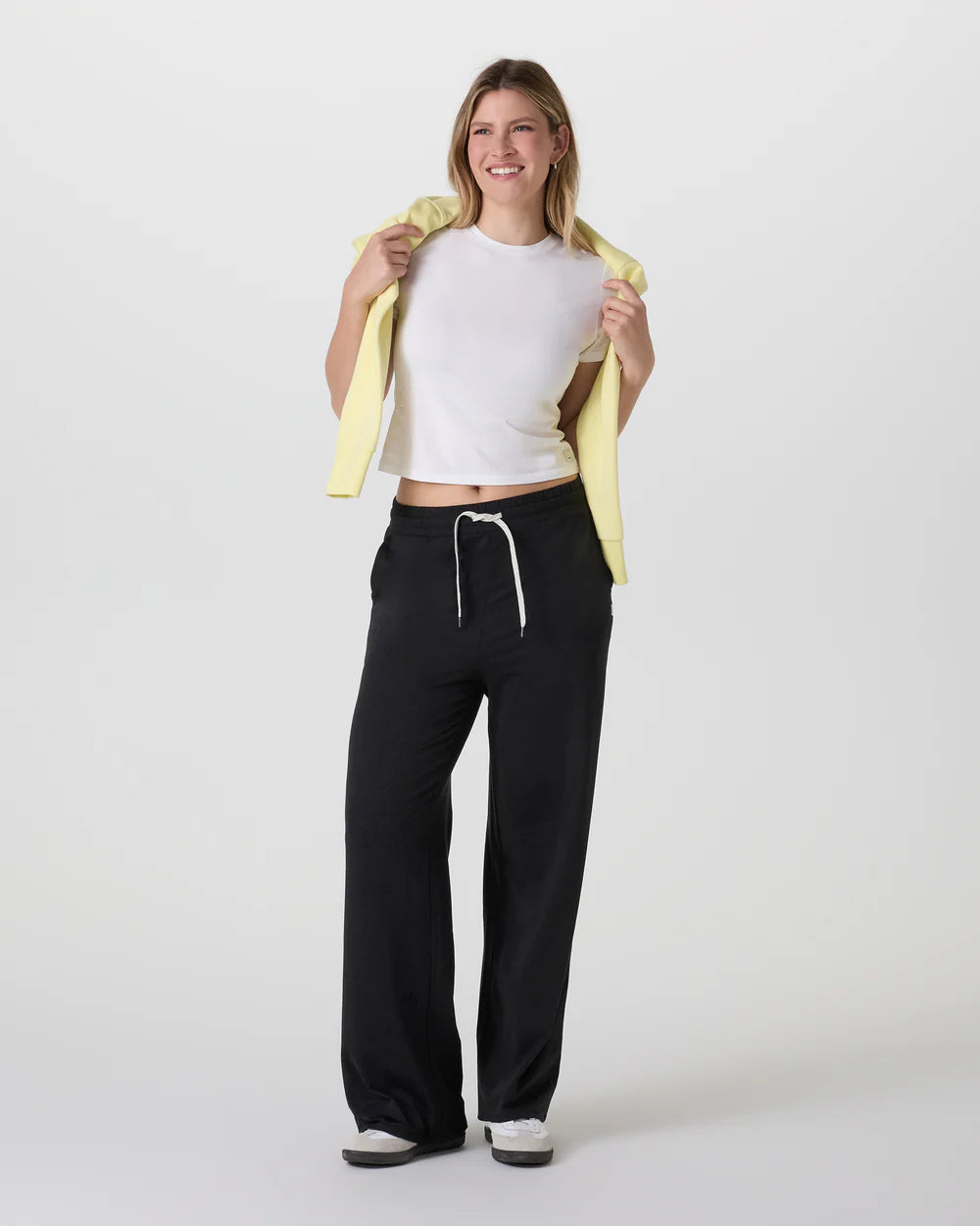 Woman wearing a white crop top and black pants with a yellow jacket over her shoulder on a white background