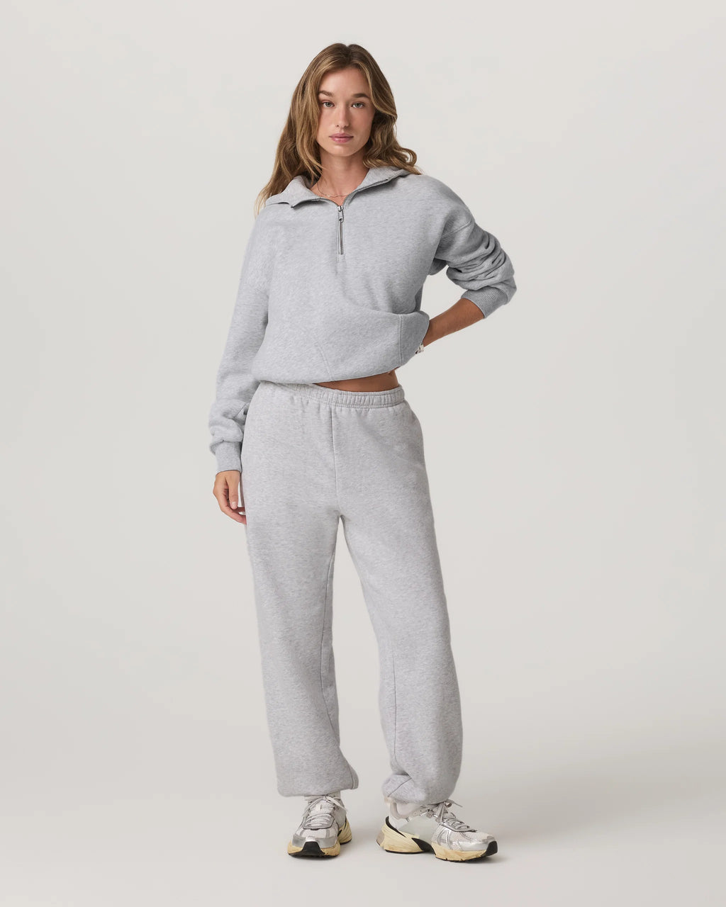 A woman modeling a light gray heathered two-piece loungewear set consisting of a cropped half-zip sweatshirt and matching sweatpants, paired with white sneakers, against a plain light gray studio background.