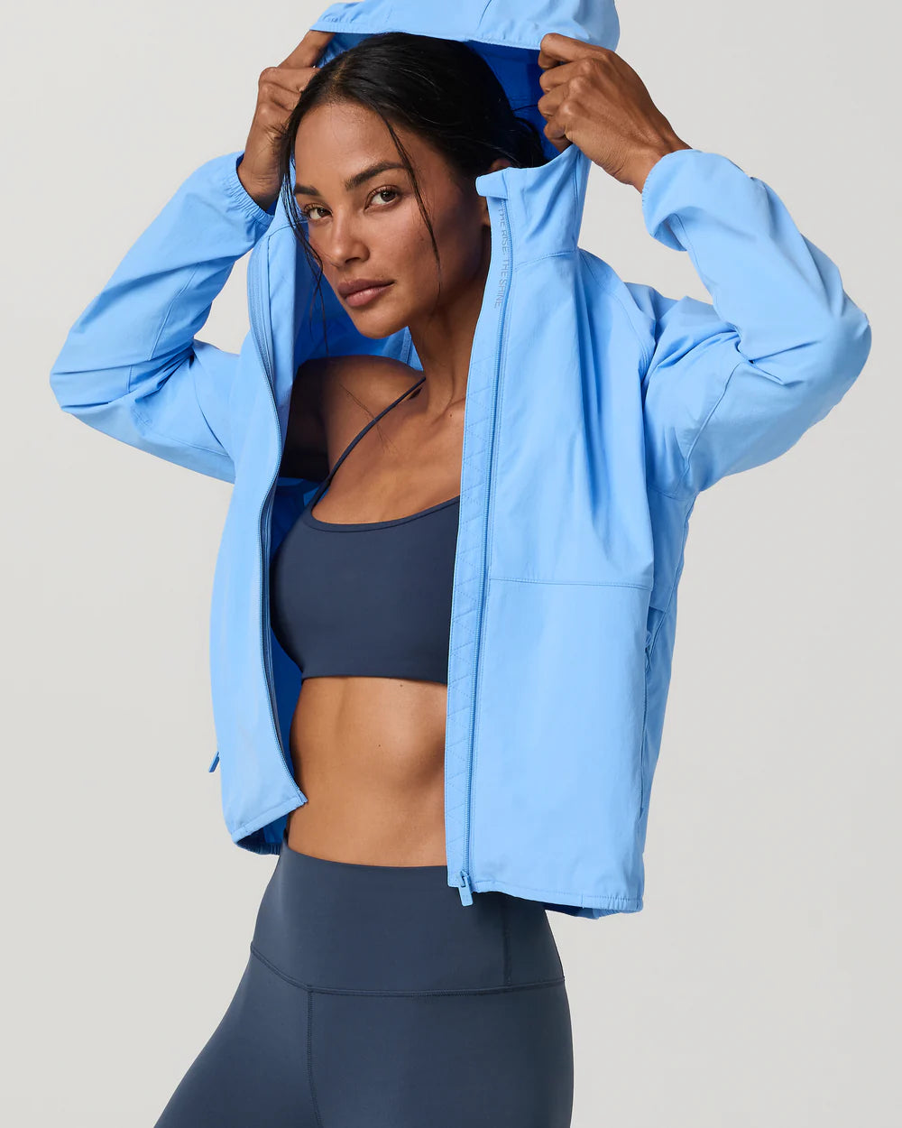 A woman wearing a light blue lightweight zip-up hooded jacket over a navy sports bra and matching leggings, posing against a light gray studio background.