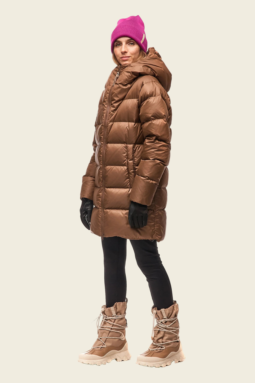 Person wearing a brown puffer coat, pink beanie, black gloves, and tan boots on a white background