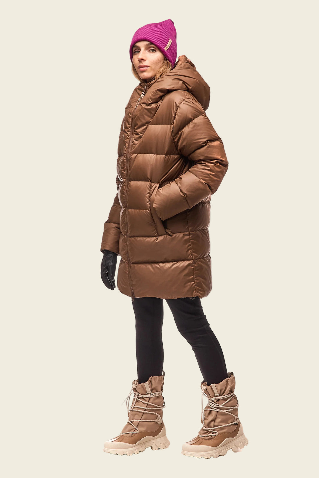 Person wearing a brown puffer coat, pink beanie, black gloves, and tan boots on a white background