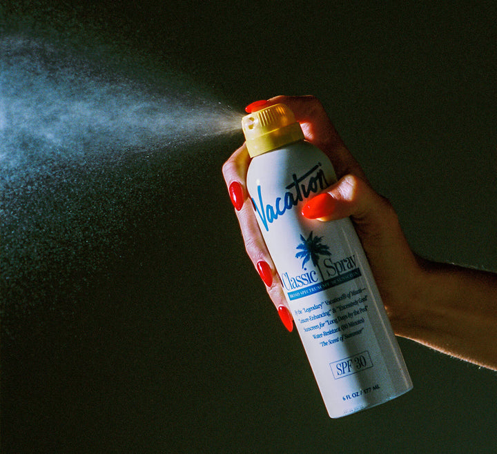Hand holding a sunscreen spray can labeled 'Vacation' against a dark background