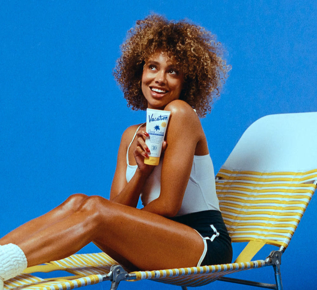 Woman sitting on a beach chair holding a sunscreen bottle against a blue background